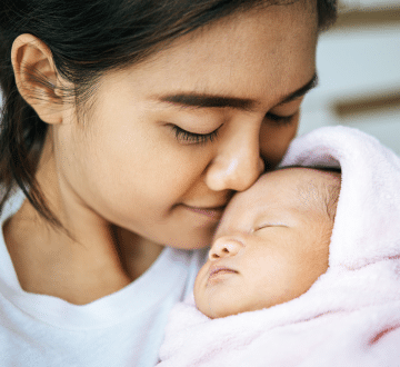 A woman gently touches her nose to a sleeping baby wrapped in a pink blanket.