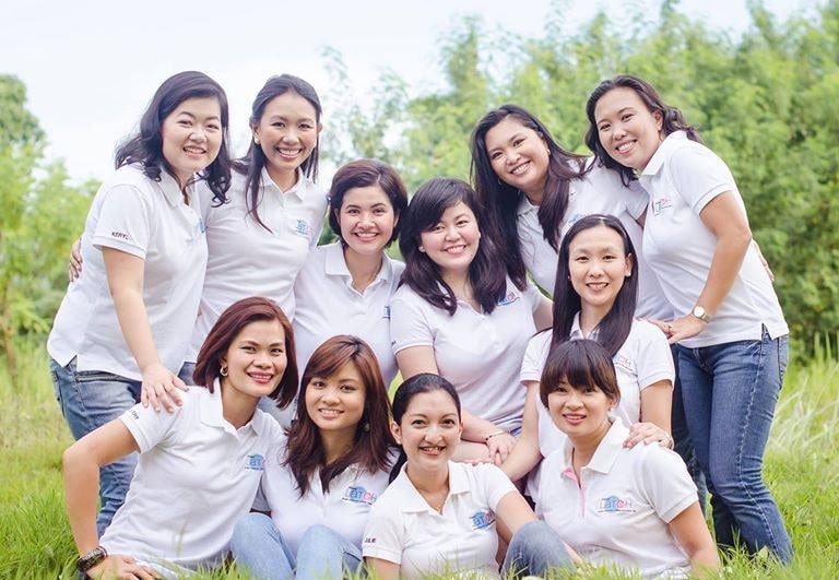 A group of ten women, all wearing white polo shirts, are smiling and posing together outdoors in a grassy area with greenery in the background.